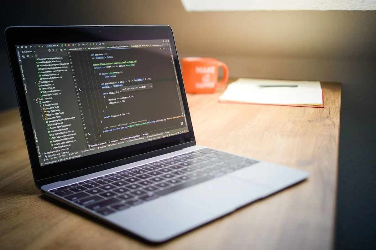 Un ordinateur portable ouvert sur du code, posé sur un bureau en bois avec un carnet et une tasse orange